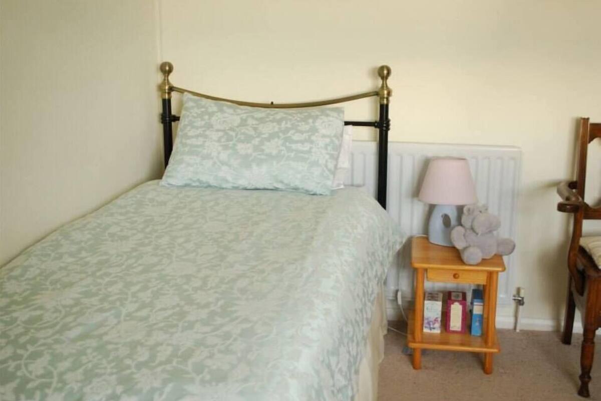 A single bed with a patterned bedspread, a bedside table with a lamp and stuffed toys, and a radiator in the background.