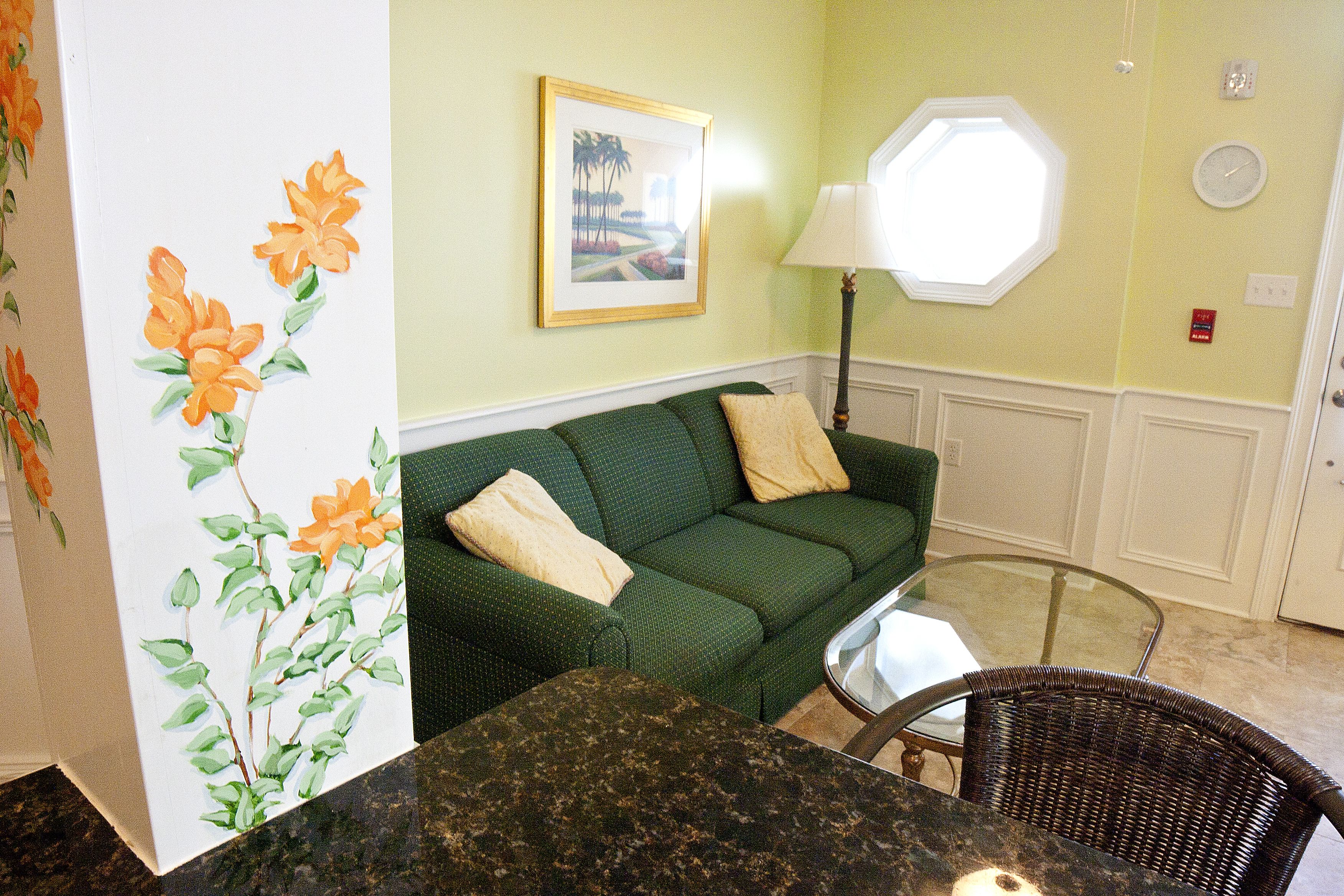 Um sofá verde com duas almofadas, uma mesa de vidro e uma cadeira de vime em um quarto com um quadro na parede e uma janela.