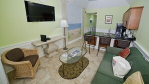 Een woonkamer met een televisie, een bank, een stoel, een salontafel en een keukenhoek met een koelkast en een spoelbak.