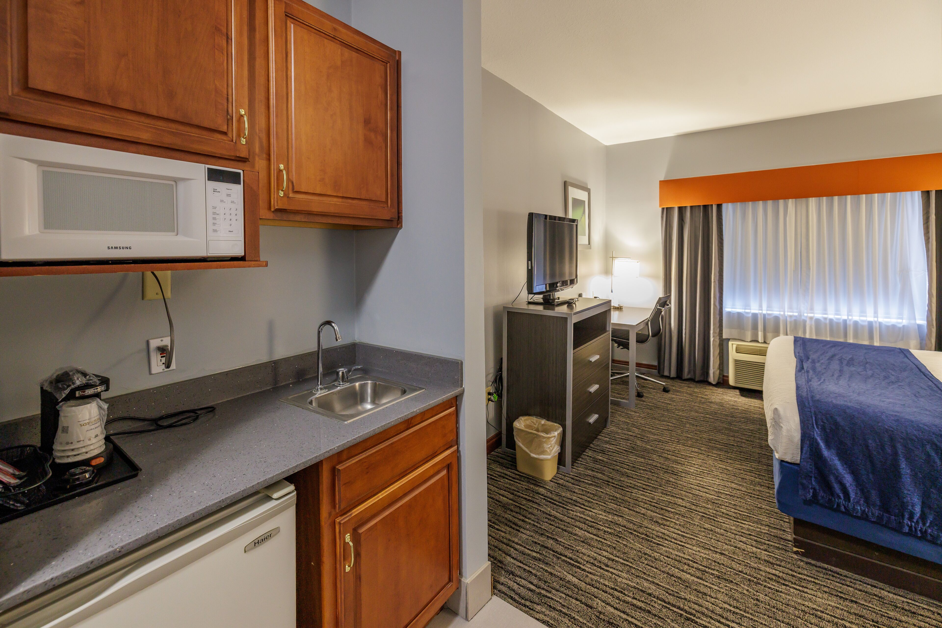 A hotel room with a kitchenette, a bed, a TV, and a window with curtains.