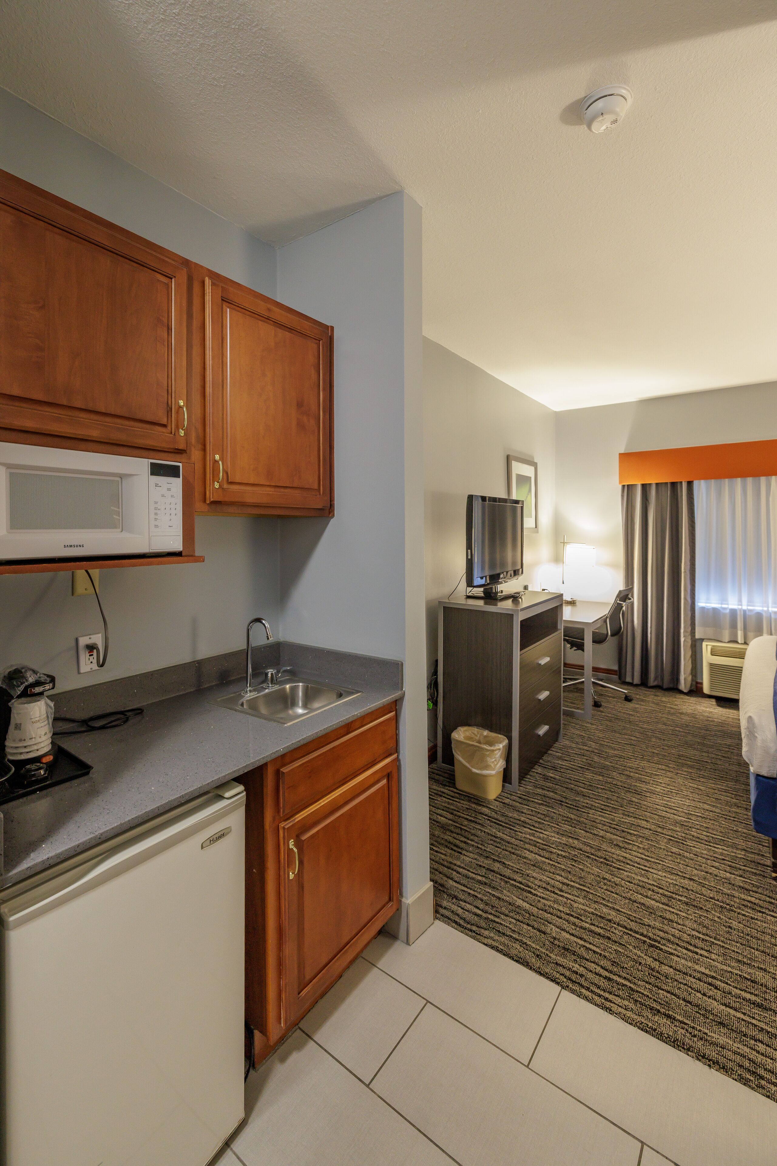 A compact hotel room with a kitchenette, a small desk, a television, and a bed with a view.