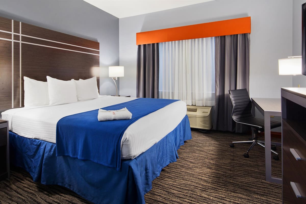 A hotel room with a large bed, two bedside tables with lamps, a desk with a chair, a television, and a window with curtains.