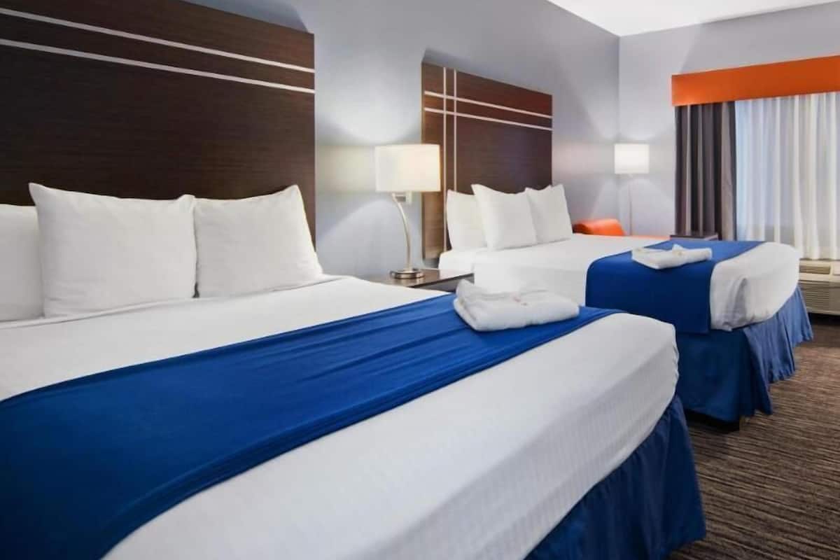 A hotel room with two beds, a nightstand, a lamp, and a window with curtains.