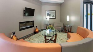 Une chambre d’hôtel moderne avec une table basse en verre, des fauteuils orange, un téléviseur à écran plat et un tableau encadré au mur.