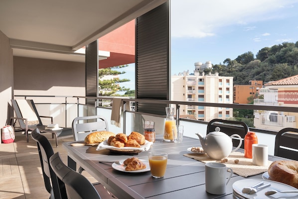 A balcony with breakfast items, including croissants, juice, and a teapot, overlooking a cityscape. - Pierre & Vacances Residence Premium Julia Augusta (Roquebrune-Cap-Martin)
