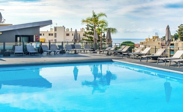 Una piscina al terrat amb gandules i ombrelles, envoltada d'edificis i palmeres.