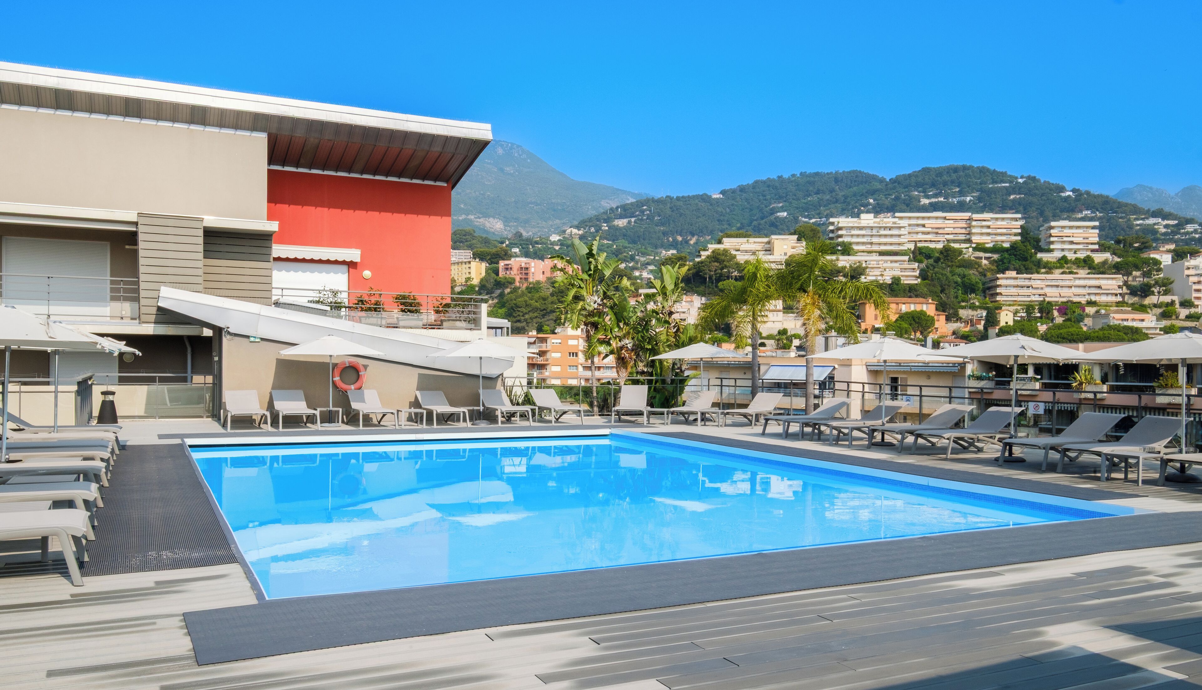 A rooftop pool with lounge chairs and umbrellas, surrounded by buildings and a mountain backdrop.