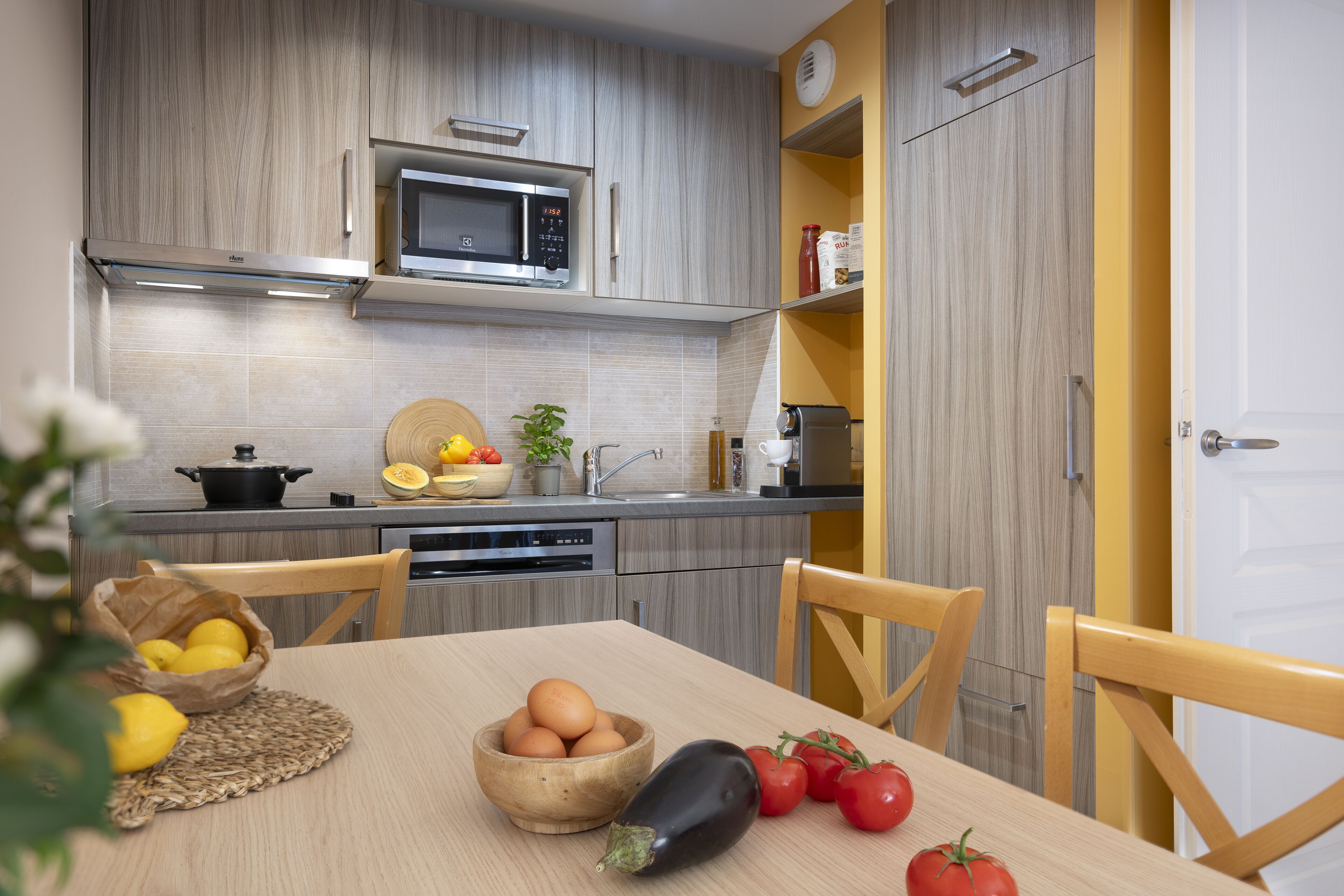 Una cocina moderna con armarios de madera, un microondas y un lavamanos. Se observa una mesa con huevos, tomates y una berenjena.