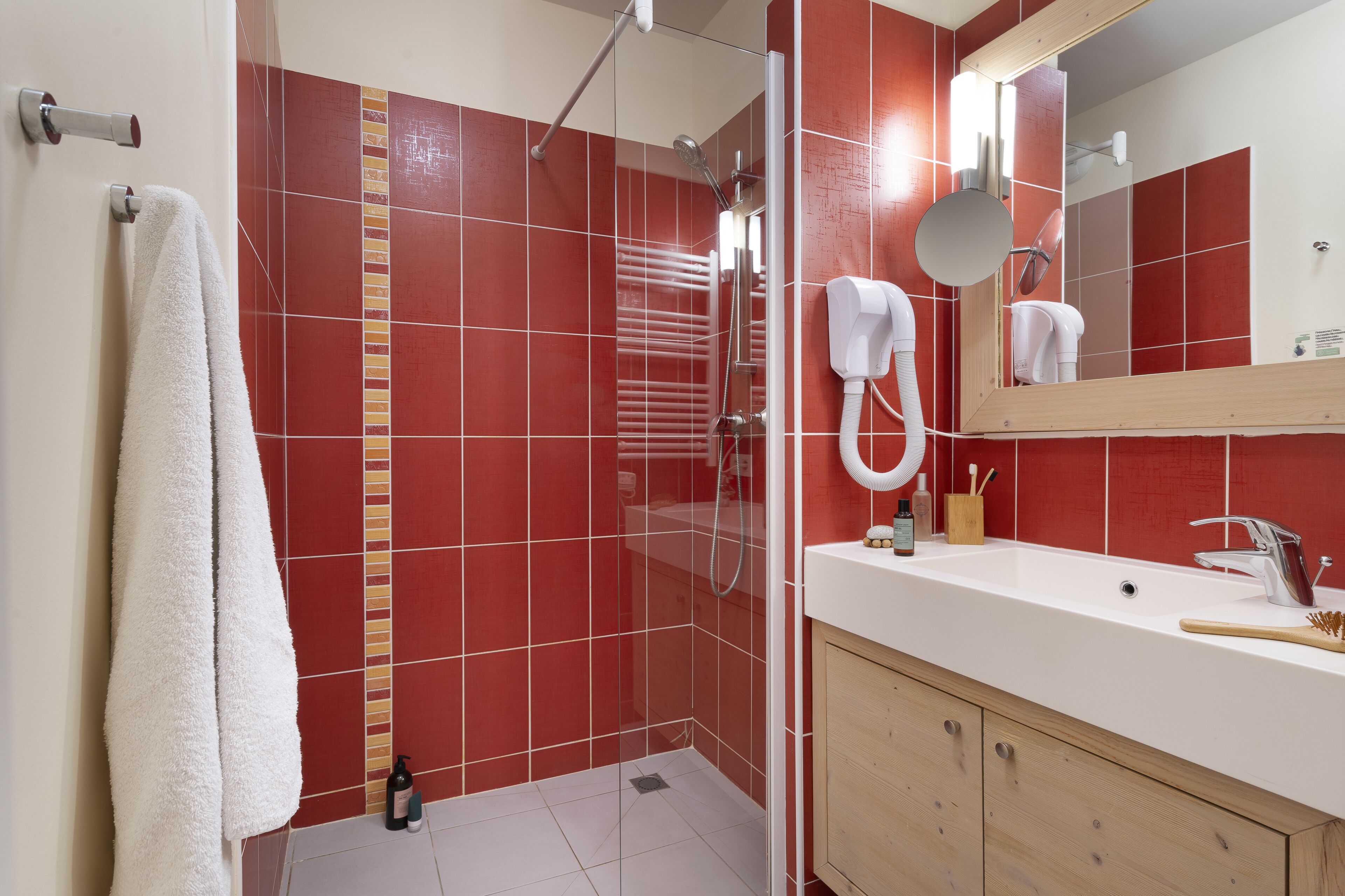 Een badkamer met rode betegelde wanden, een wit wastafelmeubel, een spiegel en een douchehoek.