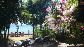 A beachside lounge with lounge chairs and a table, surrounded by lush greenery and flowering plants.