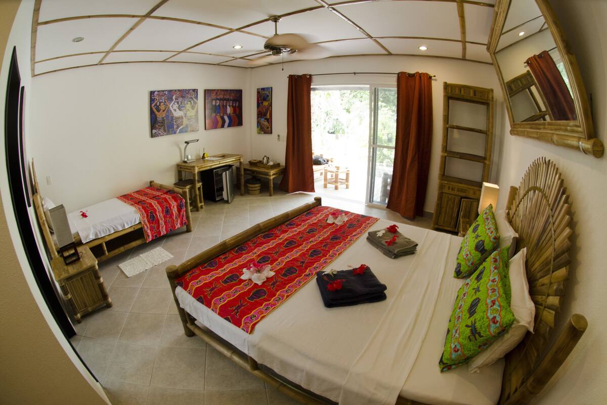 A room with a large bed, a desk, a chair, and a small table. There is a window with curtains, a mirror on the wall, and a ceiling fan.