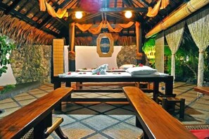 A room with a bed, wooden benches, a stone wall, and a thatched roof. - Dolphin House (Moalboal)