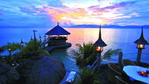 A sunset view over a calm sea with a small island structure featuring a gazebo and lanterns.