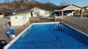 Ein Swimmingpool mit klarer, blauer Wasseroberfläche, umgeben von einem eingezäunten Bereich mit Liegestühlen und einem kleinen Gebäude mit schrägem Dach.