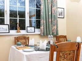 Un comedor con una mesa preparada para dos personas, con mantel blanco, una silla de madera con respaldo estampado y una ventana con cortinas florales.