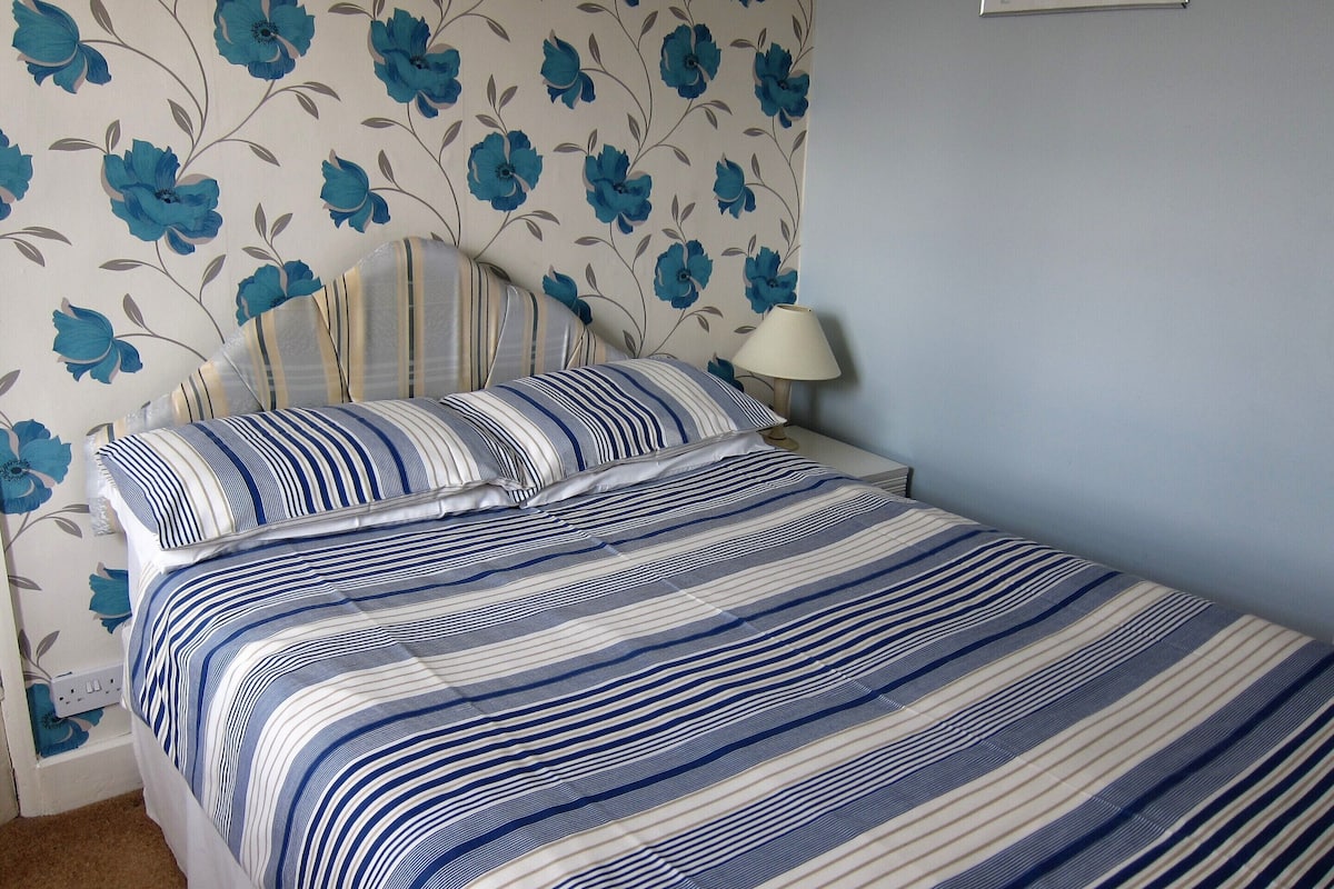 Una cama bien hecha con ropa de cama a rayas, una lámpara de noche y papel tapiz floral.