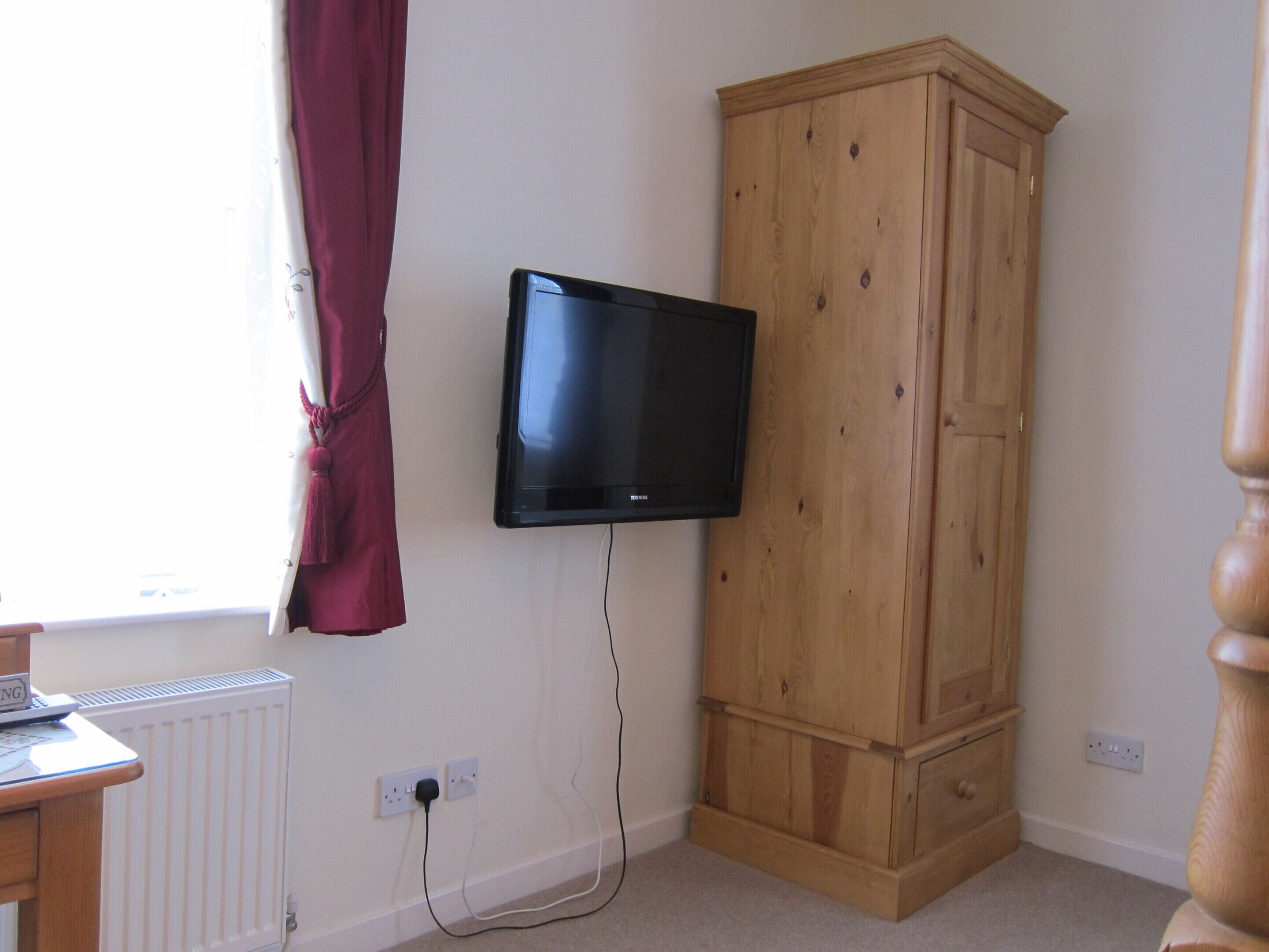 A room with a wooden wardrobe, a mounted television, a radiator, and a window with curtains.