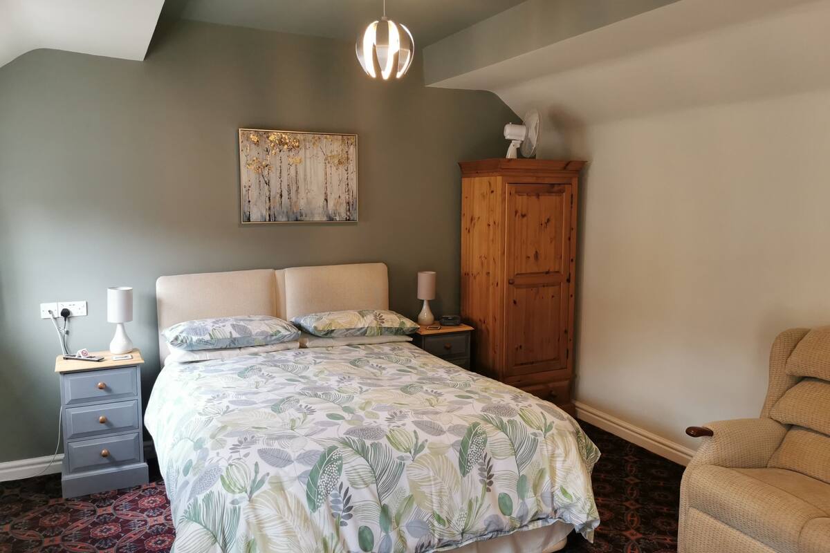 Un dormitorio con cama, mesita de noche, armario de madera, un cuadro colgado en la pared y una lámpara del techo.