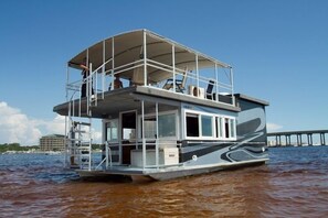 Una casa flotante con cubierta, balcón y vista a un muelle al fondo.