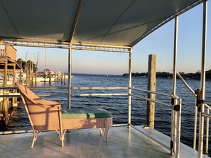 Cubierta de barco con una silla de mimbre y un asiento acolchado, con vistas a un puerto deportivo con barcos y una costa lejana.