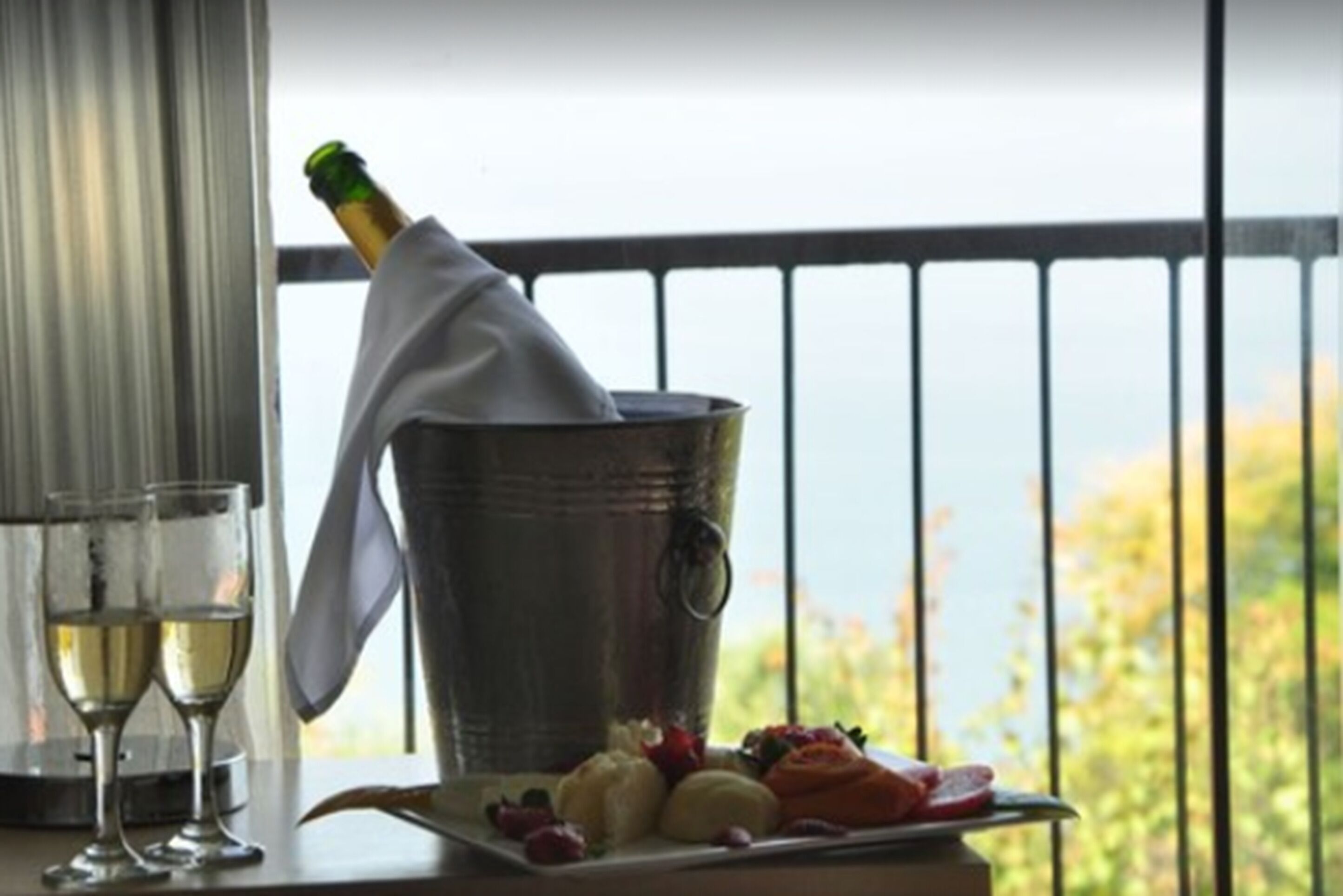 A table with a bottle of champagne, two glasses, and a fruit platter on a balcony with a railing and a view of the outdoors.