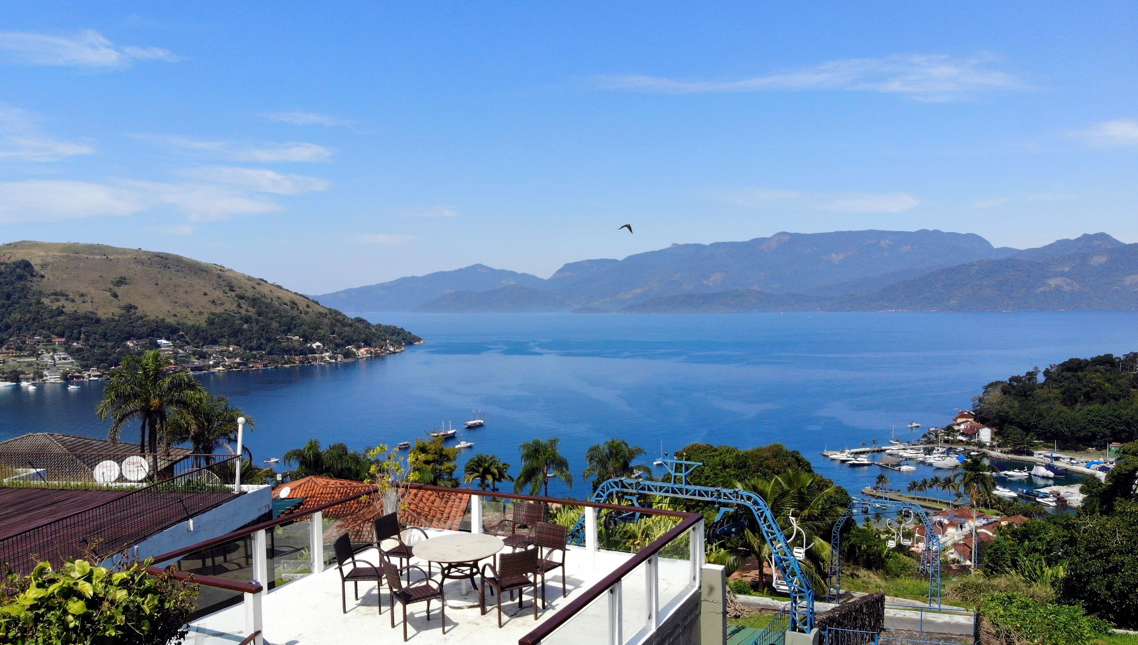 A coastal view with a chair and table on a terrace, overlooking a calm sea and distant hills.