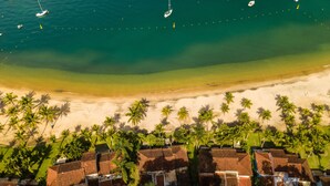 An aerial view of a beach resort with bungalow-style buildings, a sandy beach, and palm trees. - Portogalo Suite Hotel (Angra dos Reis)