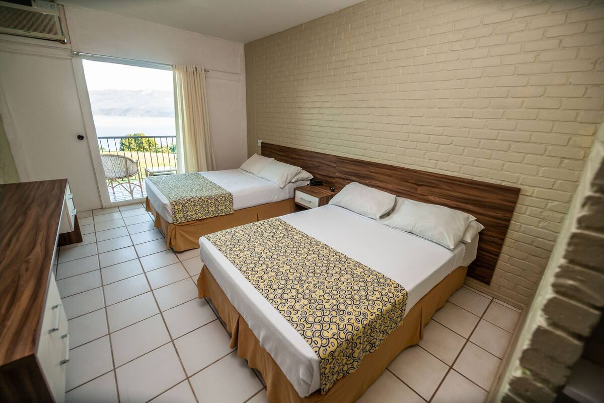 A hotel room with two beds, a wooden headboard, a balcony with a chair, and a view of the mountains.