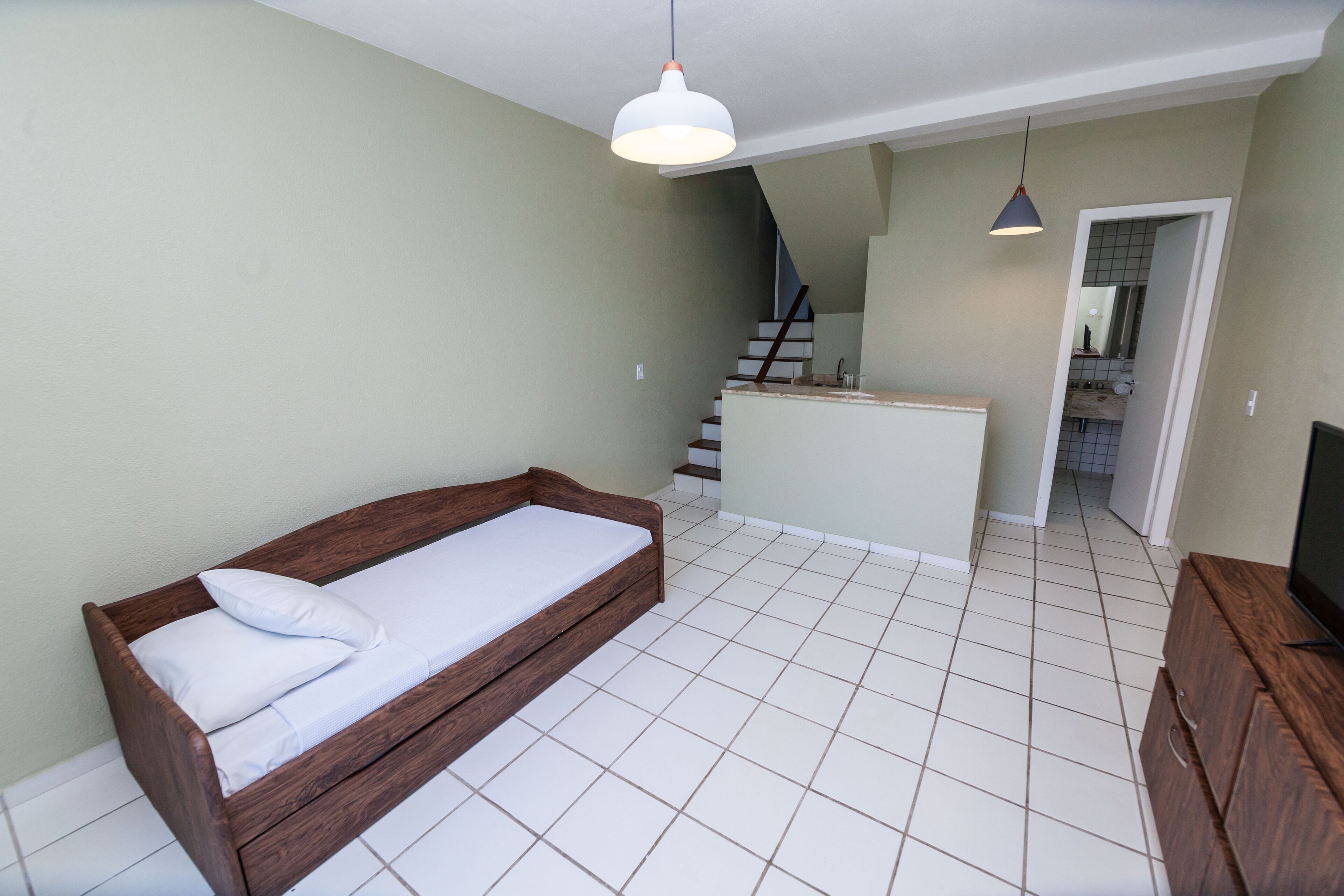 A bedroom with a wooden bed, white bedding, a small kitchenette, and a staircase leading to an upper level.