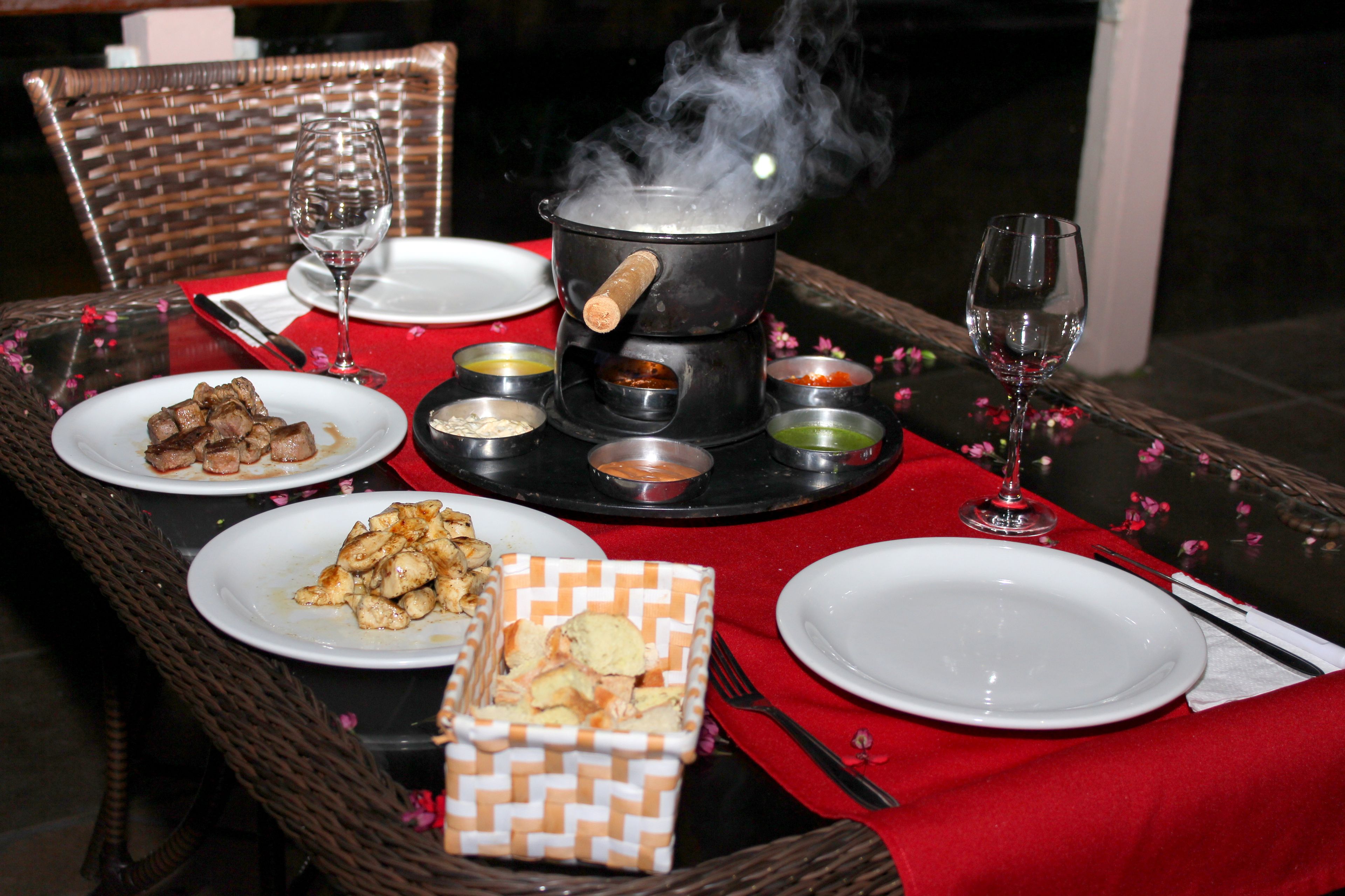 Una mesa de comedor dispuesta con platos de comida, una olla sobre un fogón portátil y diferentes condimentos.