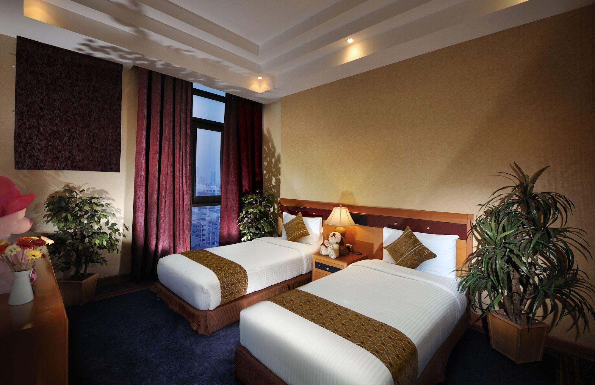 Ein Hotelzimmer mit zwei Betten, einem Nachttisch mit Lampe, einem Fenster mit Vorhängen und Topfpflanzen.