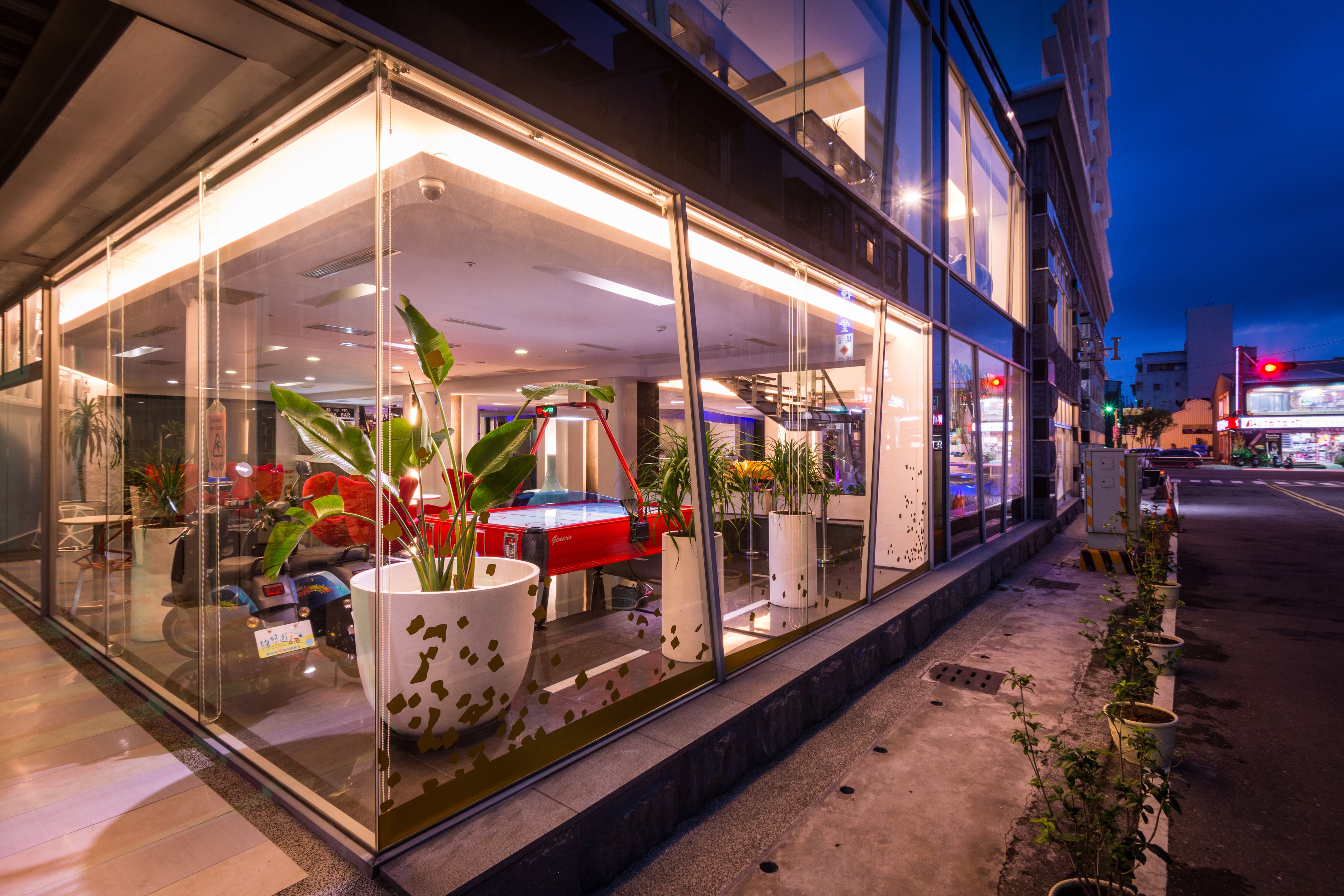 Un edificio moderno con amplios ventanales de vidrio, plantas en macetas y un auto rojo en el interior.