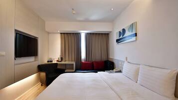 A hotel room with a large bed, a flat-screen TV mounted on the wall, a desk with a chair, a window with curtains, and a painting on the wall.