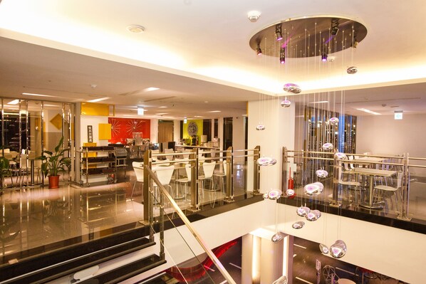 A modern hotel lobby with a glass staircase, a reception desk, and a dining area with chairs and tables.