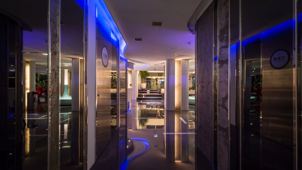 A modern hotel corridor with reflective floors and glass walls, leading to a reception area with a plant and seating. - Hotel Hi Xinmin (Chiayi City)