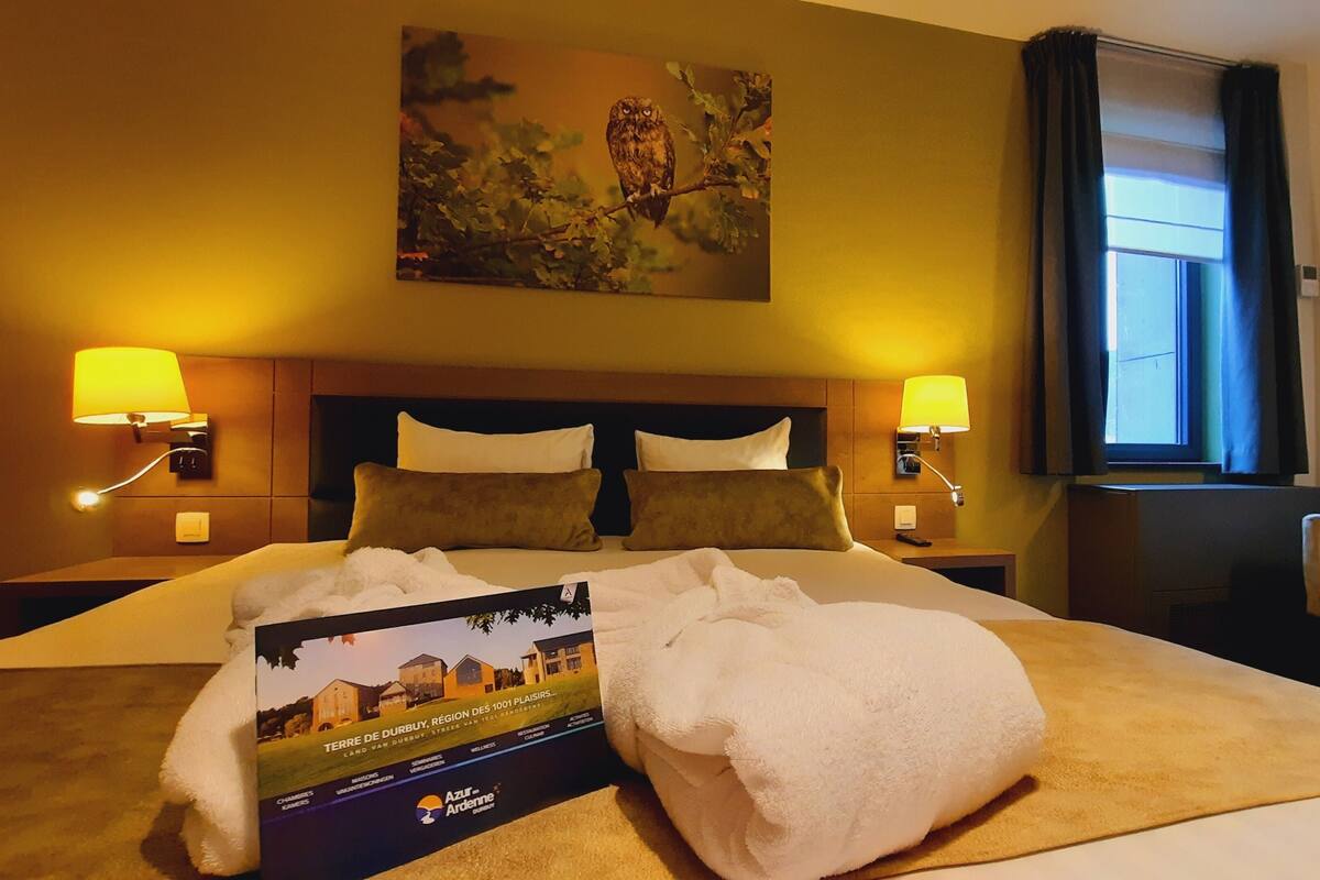 Ein Hotelzimmer mit einem großen Bett, zwei Nachttischlampen, einem Bild an der Wand, einem Fenster mit Vorhängen und einer Broschüre auf dem Bett.