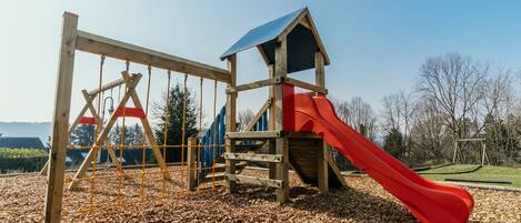 A wooden play structure with a red slide and a climbing area, set on a mulch-covered ground.
