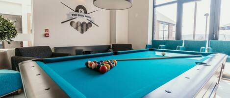 A pool table with balls, a clock, and a sign indicating 'The Pool Room' in a well-lit indoor space with large windows.