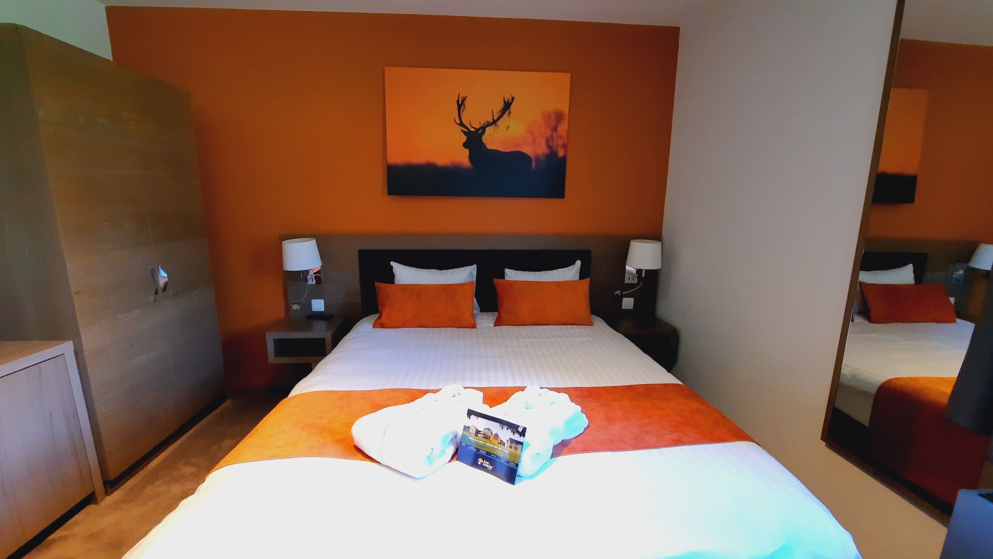 Una habitación de hotel con una cama, dos mesitas de noche con lámparas, una mesita auxiliar y un espejo grande, además de un cuadro de un ciervo en la pared.
