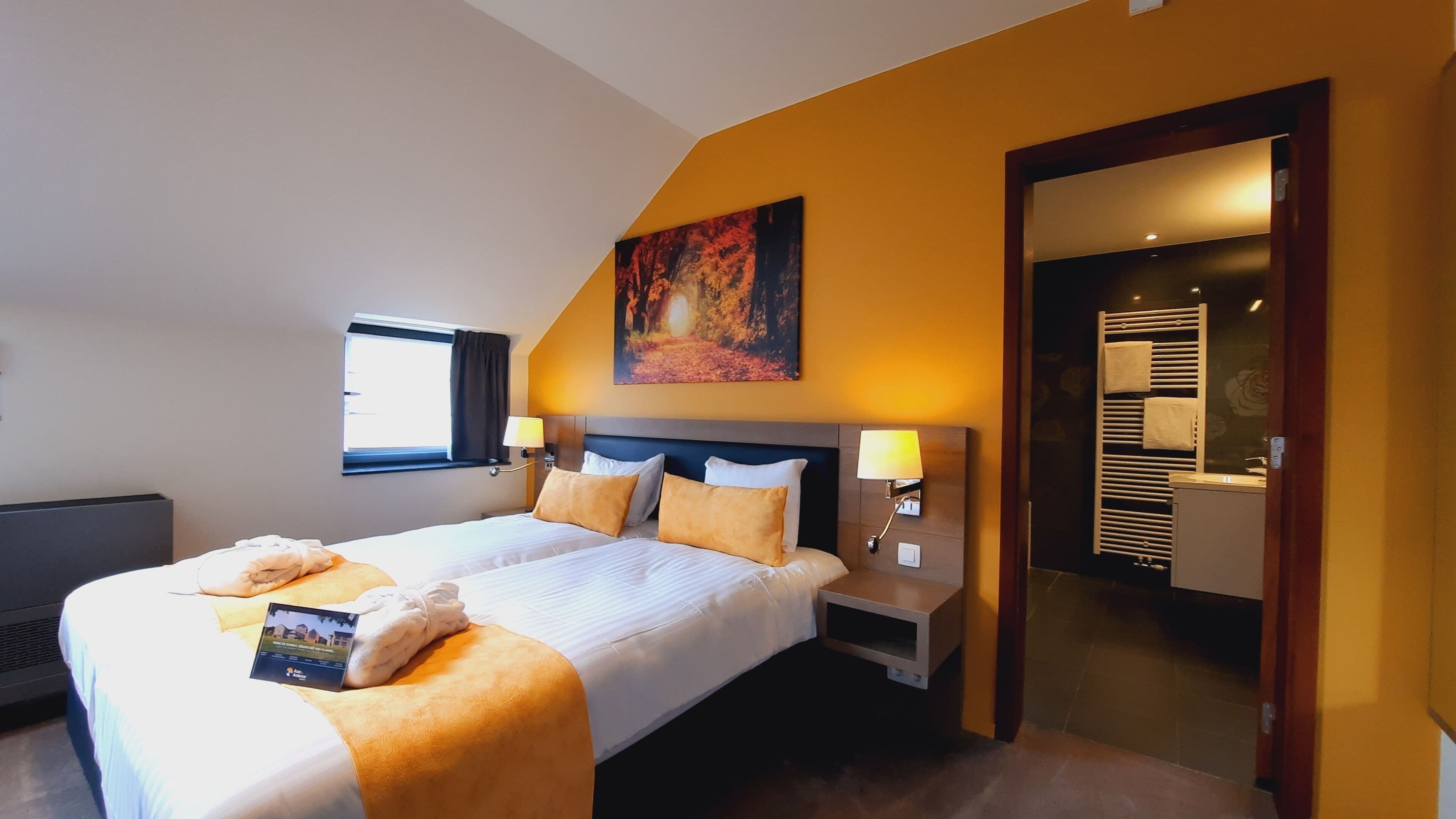 Une chambre d’hôtel avec un grand lit, deux tables de chevet, une fenêtre avec des rideaux, un tableau au mur et une salle de bain visible à travers une porte ouverte.