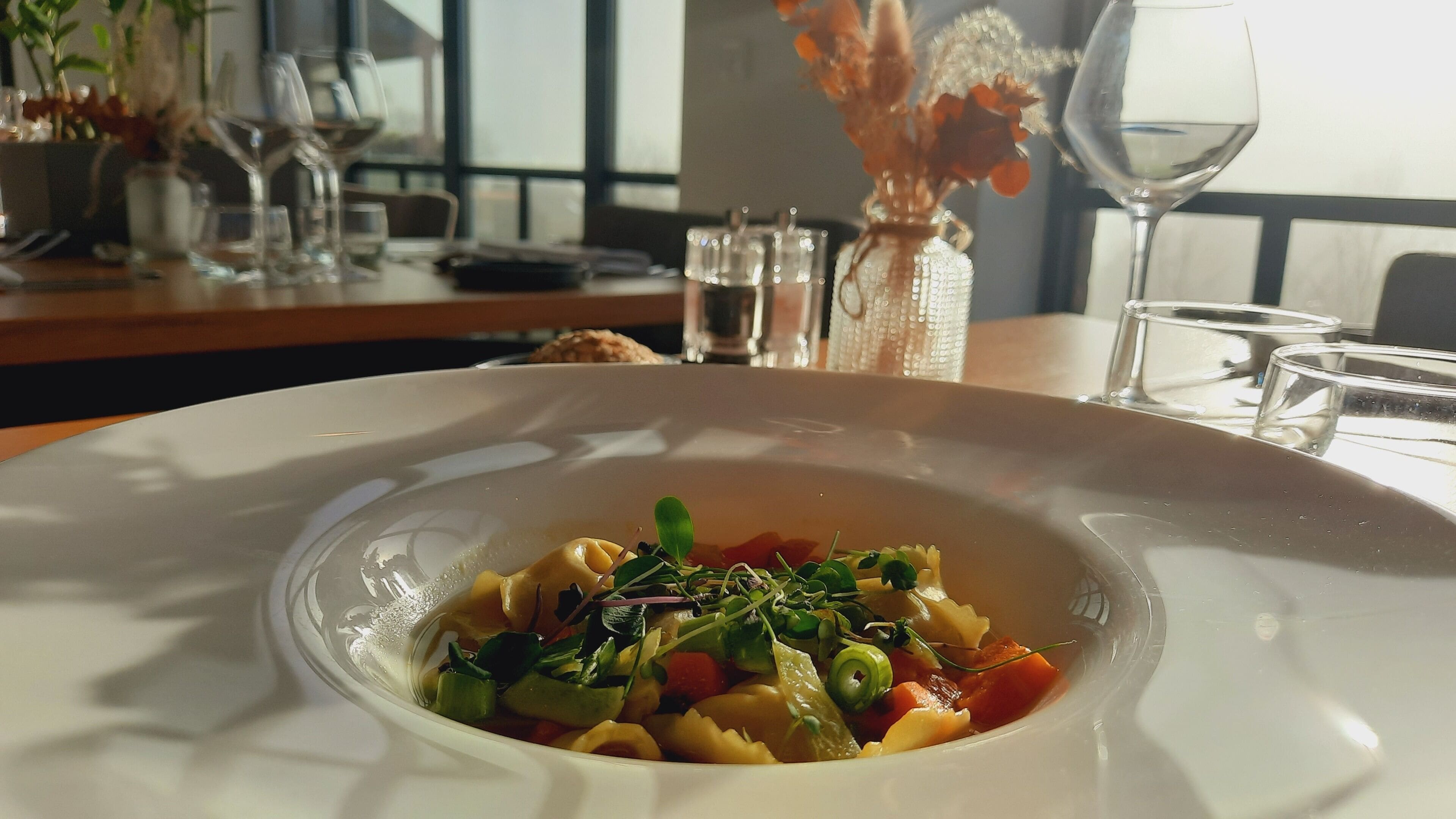 Een eettafel met een pastasalade, een vaas met gedroogde bloemen en een glas water.