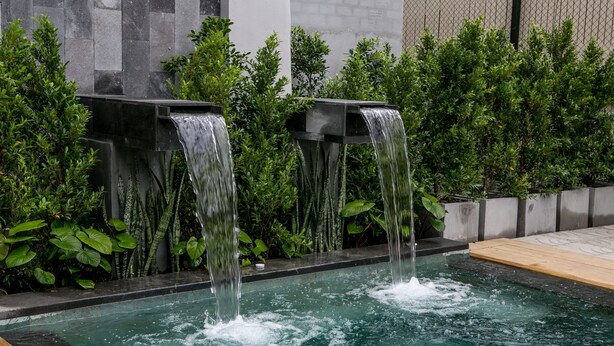 A pool with two waterfalls, surrounded by lush greenery and a stone wall. - Amenity Apartel Samui (Koh Samui)