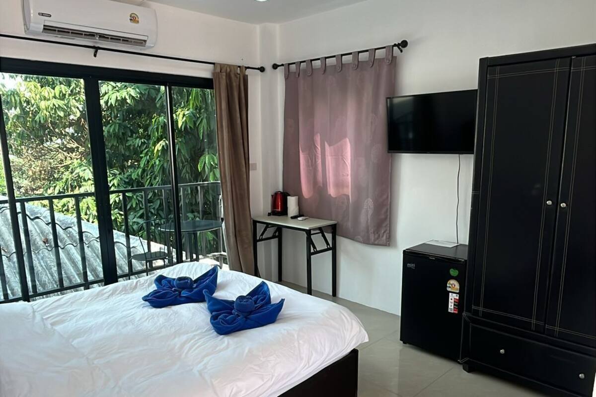 A hotel room with a large bed, an air conditioning unit, a TV, a wardrobe, and a balcony with a view of greenery.