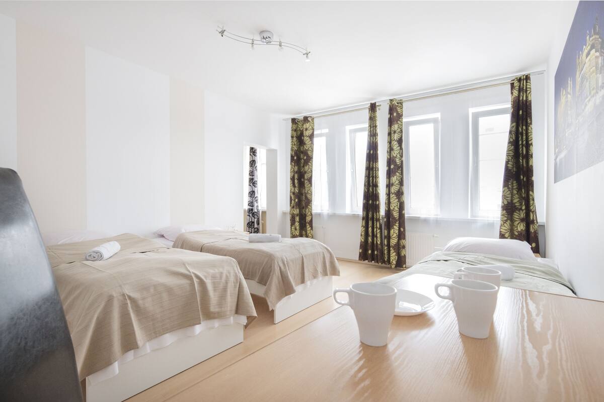 A hotel room with two beds, a wooden table with two white mugs, and a large window with curtains.