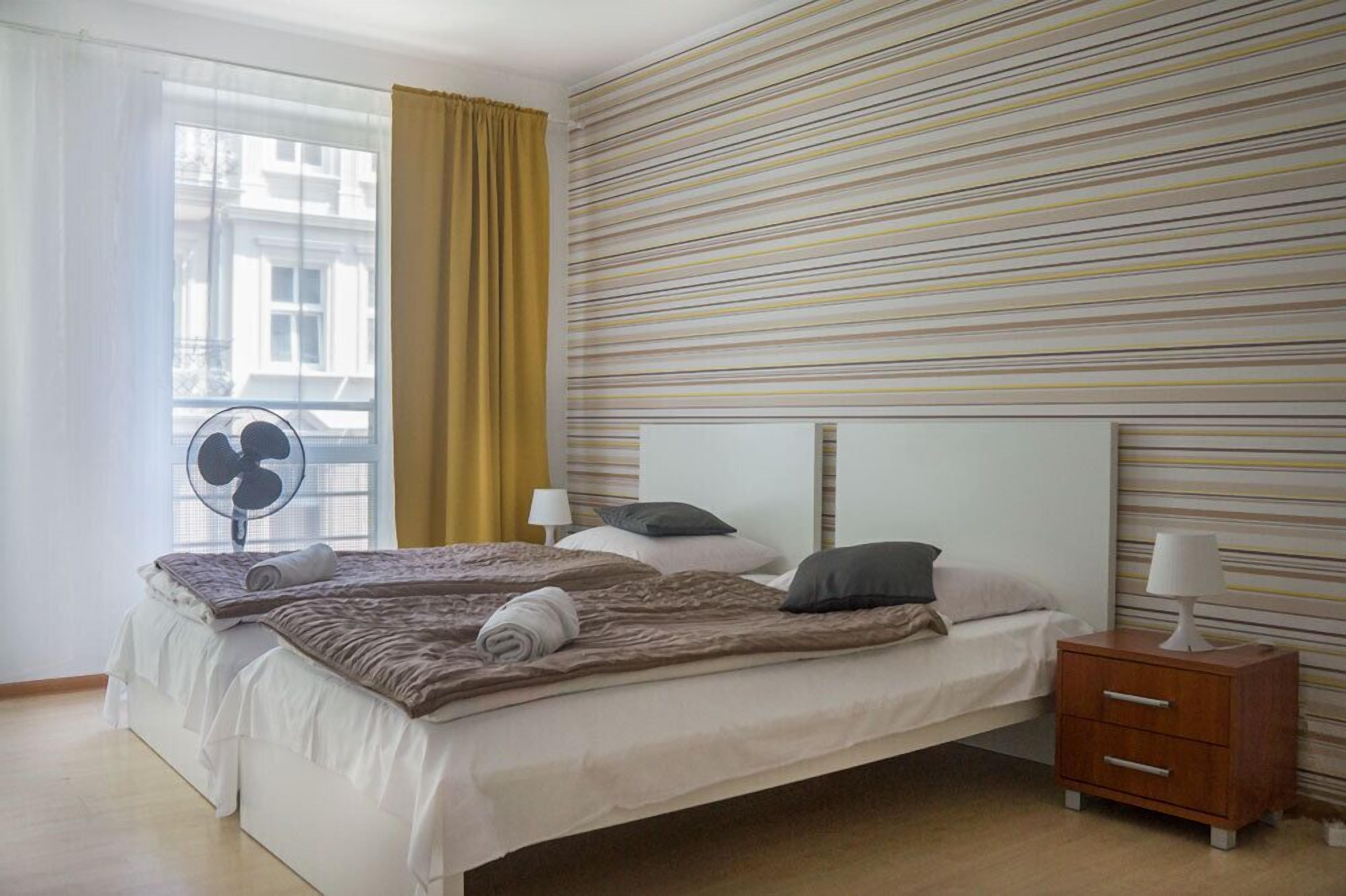 Ett hotellrum med en stor säng, ett nattduksbord med en lampa, en fläkt och ett fönster med gardiner.