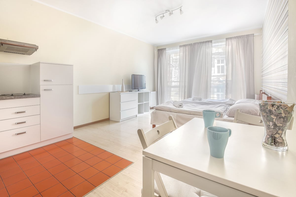 A modern, well-lit room with a kitchenette, a dining area with a table and chairs, a bed with a view, and a large window with curtains.