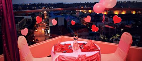 A table set for two with a city view, red tablecloth, and heart-shaped balloons.