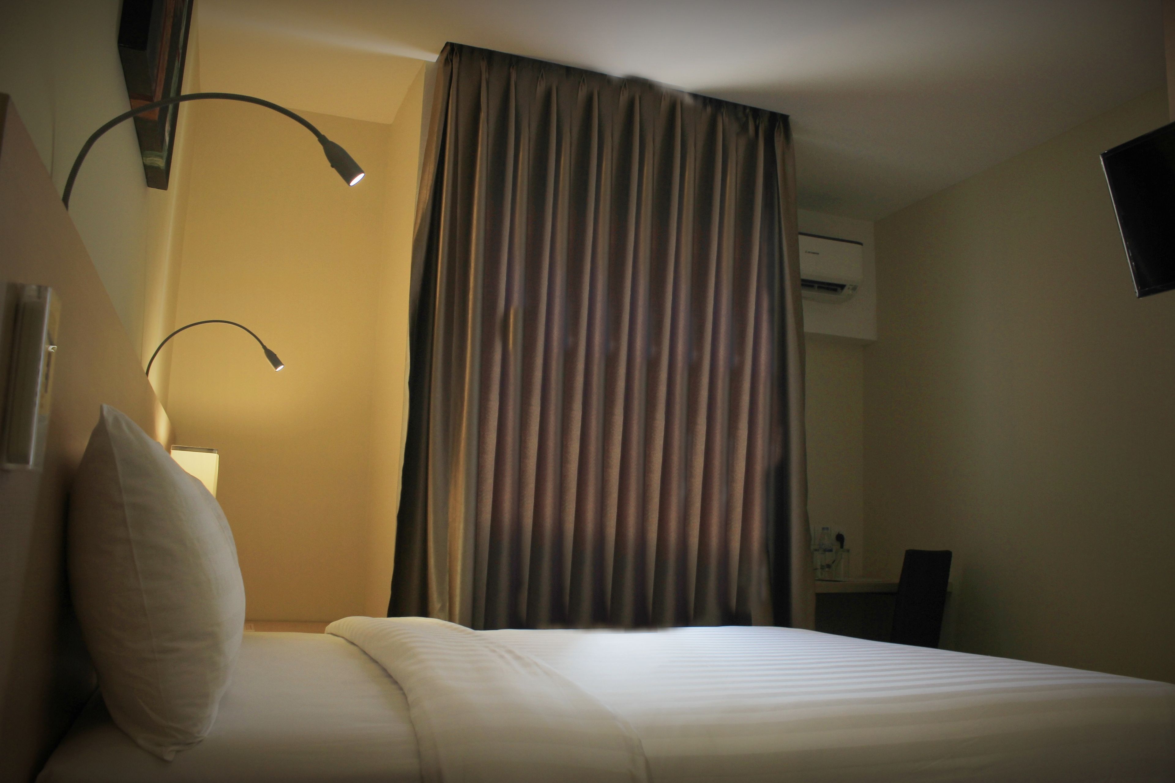 A hotel room with a bed, a nightstand, a lamp, a curtain, and an air conditioning unit.