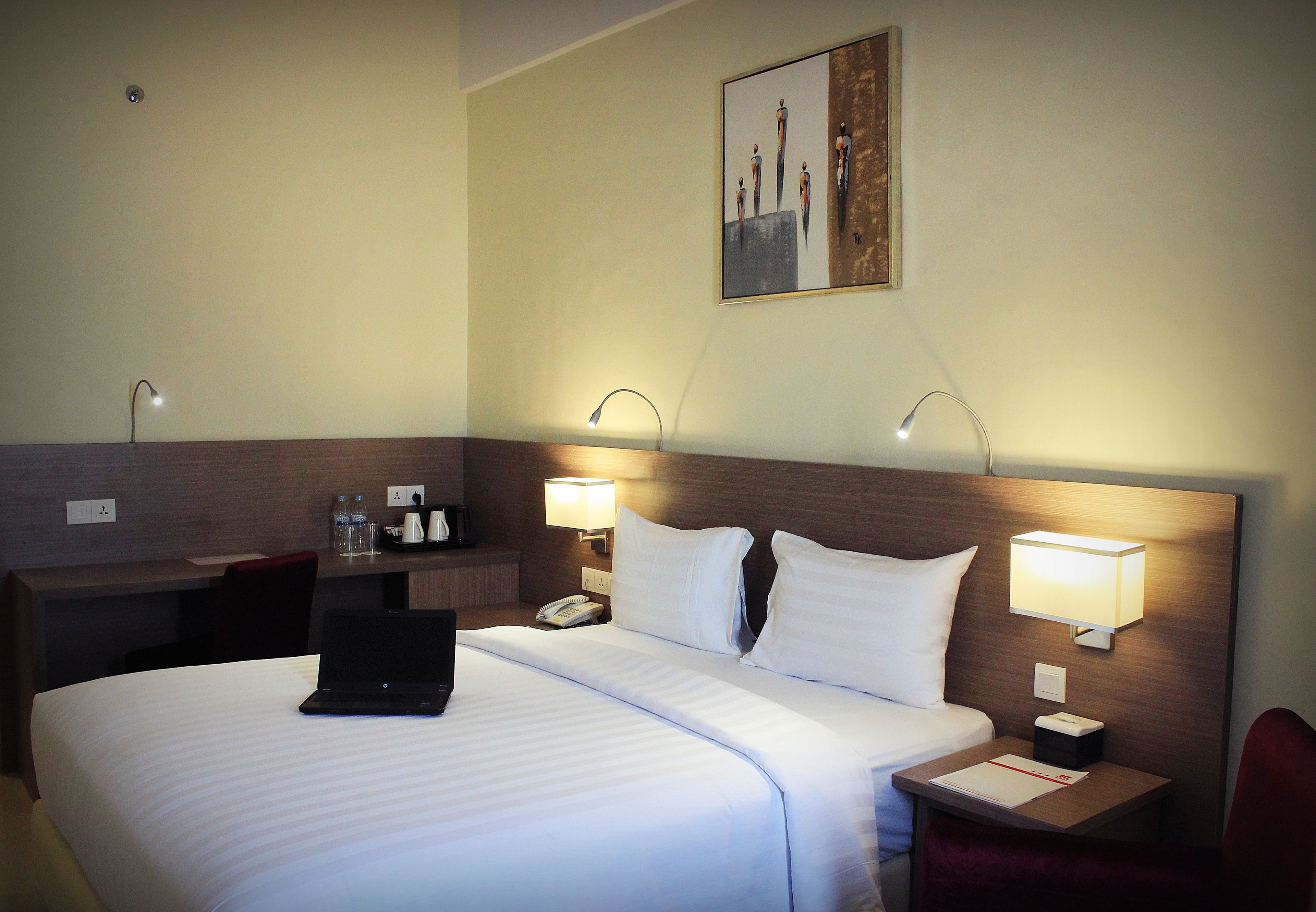 A hotel room with a neatly made bed, a desk with a laptop, a telephone, and two wall-mounted lamps.