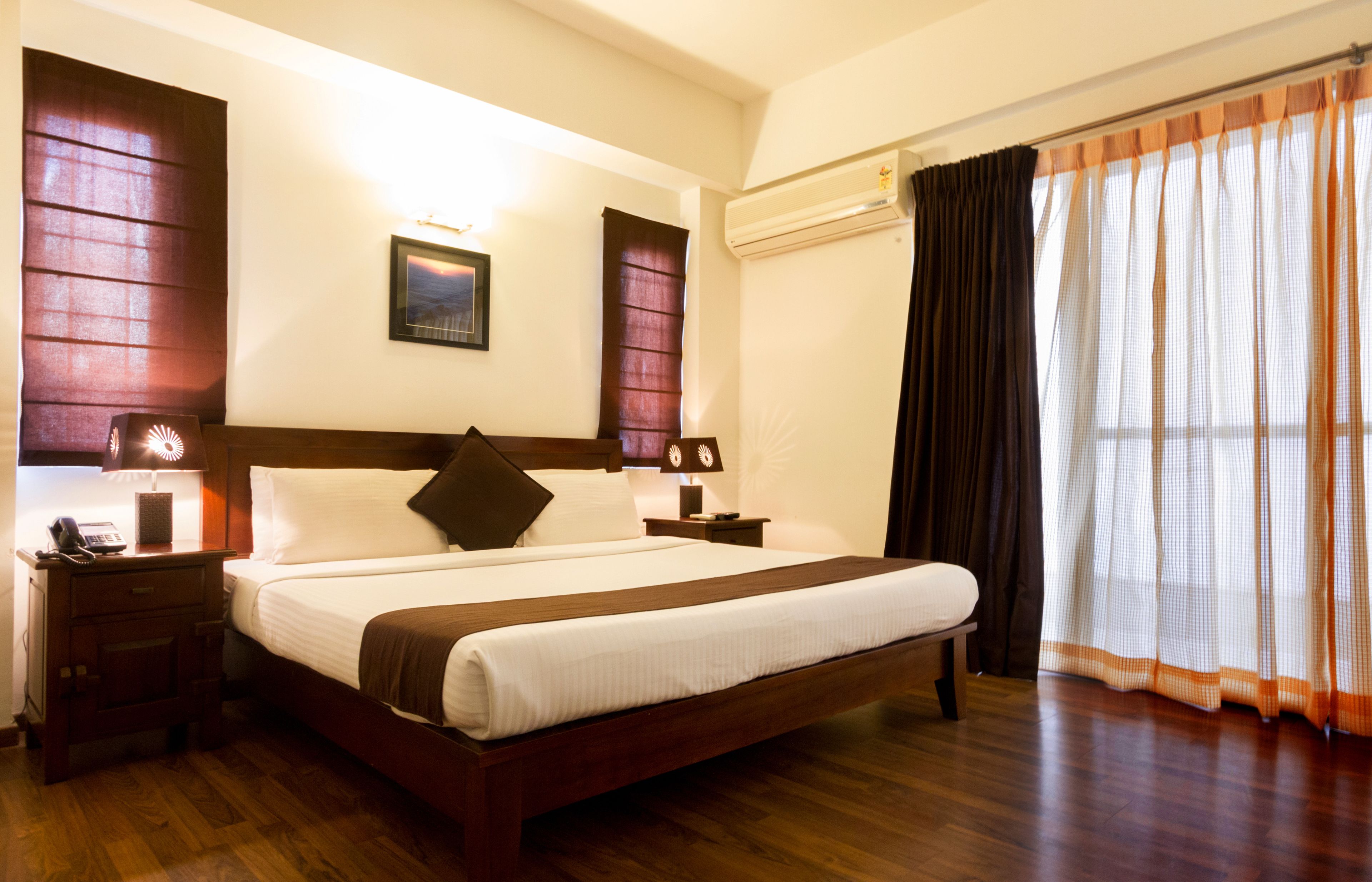 A hotel room with a large bed, wooden headboard, two bedside tables with lamps, a nightstand, and a window with curtains.